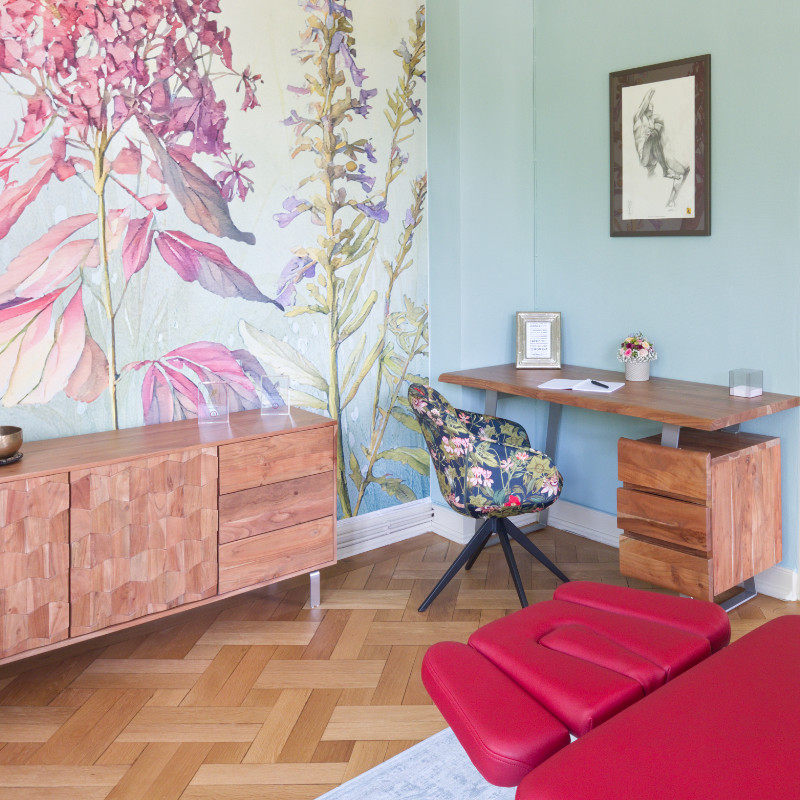 Ein heller Therapieraum mit einer roten Behandlungsliege, einem Holztisch mit Stuhl und einem Sideboard vor einer Wand mit einer floralen Tapete. Das Zimmer hat einen Fischgrät-Parkettboden.