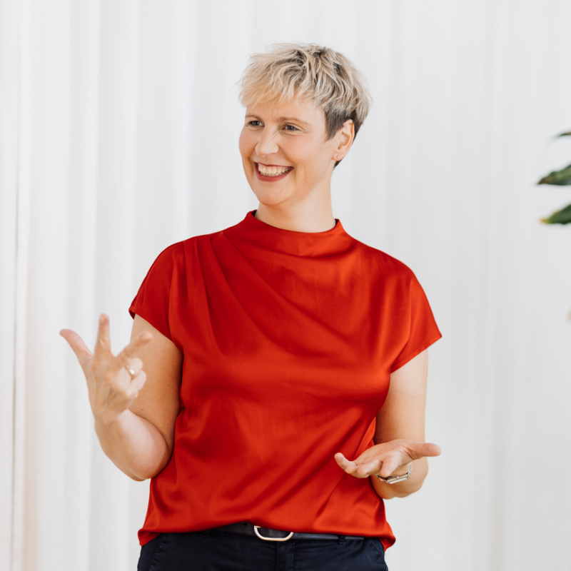 Sabine Nofz, eine lächelnde Frau mit kurzen blonden Haaren und einer roten Bluse, hält einen Vortrag und gestikuliert mit der Hand. Im Hintergrund sind helle, weiße Vorhänge zu sehen.